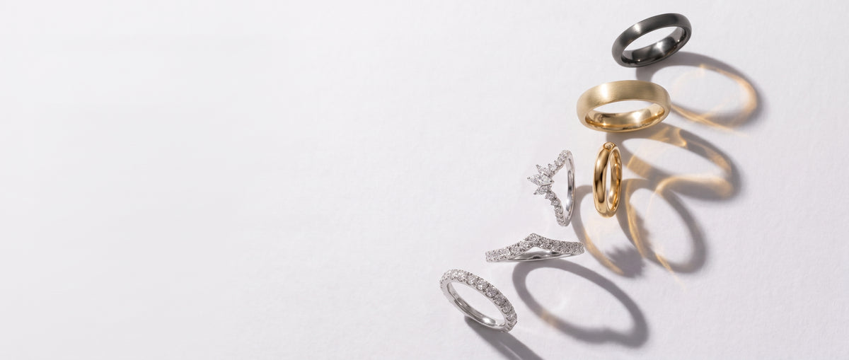 Five minimalist rings in silver, yellow gold, and black metal, arranged vertically with soft shadows on a clean white background.